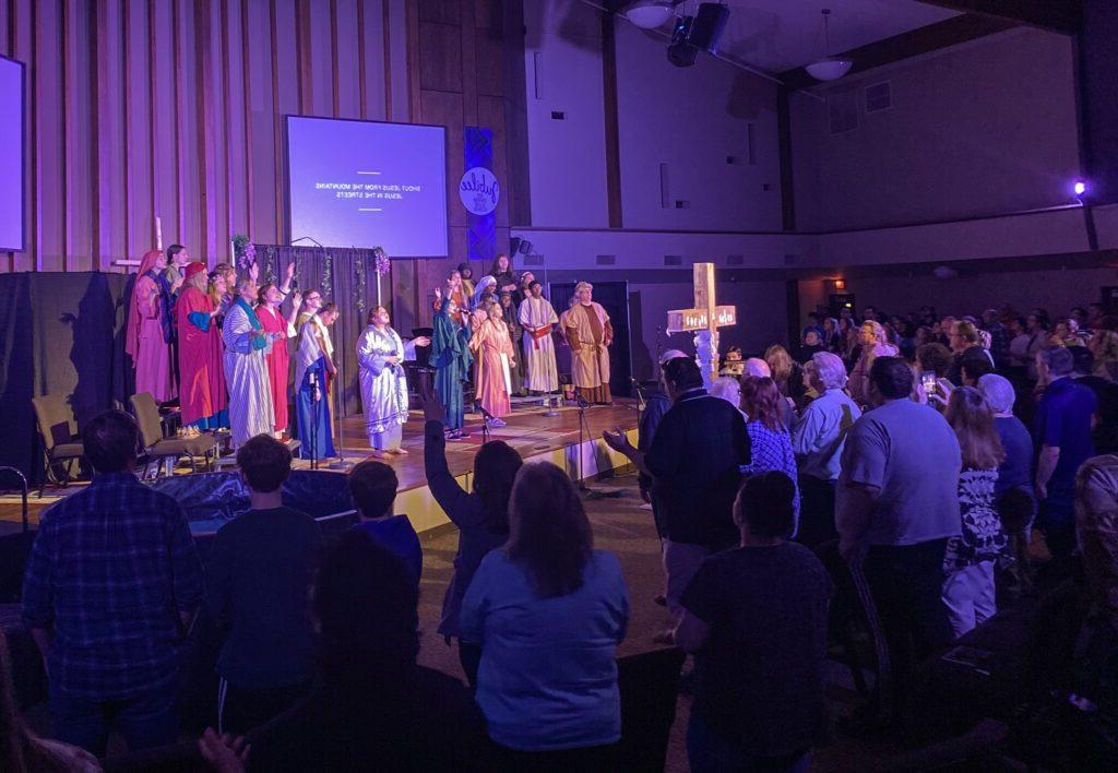 一群人, dressed in costumes, perform on a stage in front of an audience inside a church. A wooden cross stands prominently at the forefront, partially obscuring stage view.